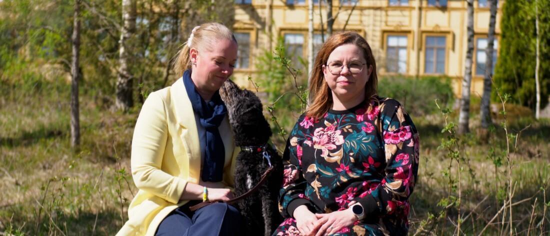 Kuvassa on kaksi naista ja musta koira istumassa puunrunkoon tehdyllä penkillä ulkona. Taustalla näkyy keltainen rakennus ja puita.