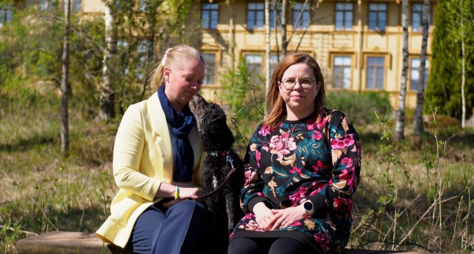 Kuvassa on kaksi naista ja musta koira istumassa puunrunkoon tehdyllä penkillä ulkona. Taustalla näkyy keltainen rakennus ja puita.