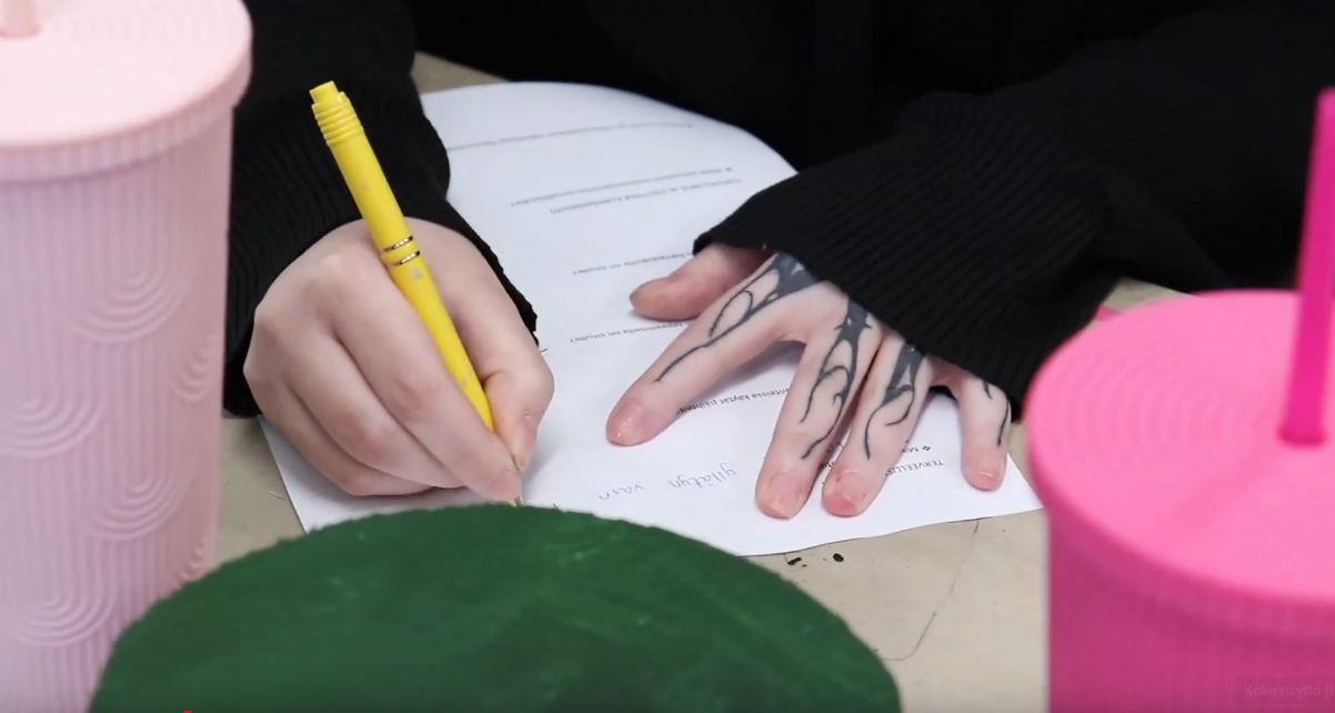 Lähikuva henkilön käsistä, jotka kirjoittavat paperille keltaisella kynällä. Oikeassa kädessä on sormissa tatuointeja.