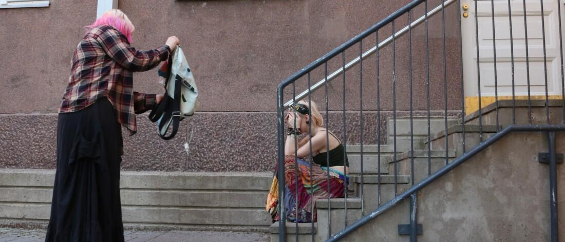 Rakennuksen ulkoportailla kaksi nuorta. Toinen seisoo ja kaivaa tavaroita repusta, toinen istuu portailla kumartuneena kädet kasvoilla.