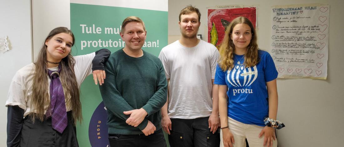 Neljä ihmistä seisoo vierekkäin sisätiloissa. Heidän takanaan on vihreä roll-up-mainos ja oikealla seinällä on käsinkirjoitettu juliste, jonka reunoille on piirretty sydämiä.