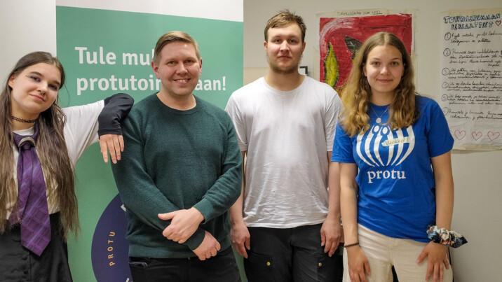 Neljä ihmistä seisoo vierekkäin sisätiloissa. Heidän takanaan on vihreä roll-up-mainos ja oikealla seinällä on käsinkirjoitettu juliste, jonka reunoille on piirretty sydämiä.