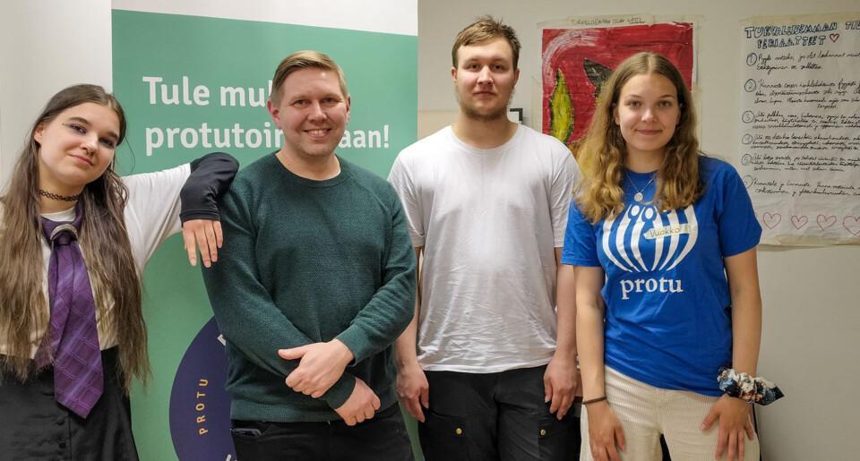 Neljä ihmistä seisoo vierekkäin sisätiloissa. Heidän takanaan on vihreä roll-up-mainos ja oikealla seinällä on käsinkirjoitettu juliste, jonka reunoille on piirretty sydämiä.