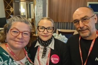 Kolme hymyilevää henkilöä yhteiskuvassa sisätiloissa. Heillä kaikilla on kaulassaan punaiset SOSTE-kaulanauhat, ja keskellä olevalla henkilöllä on lisäksi pinkki SOSTE-rintamerkki.